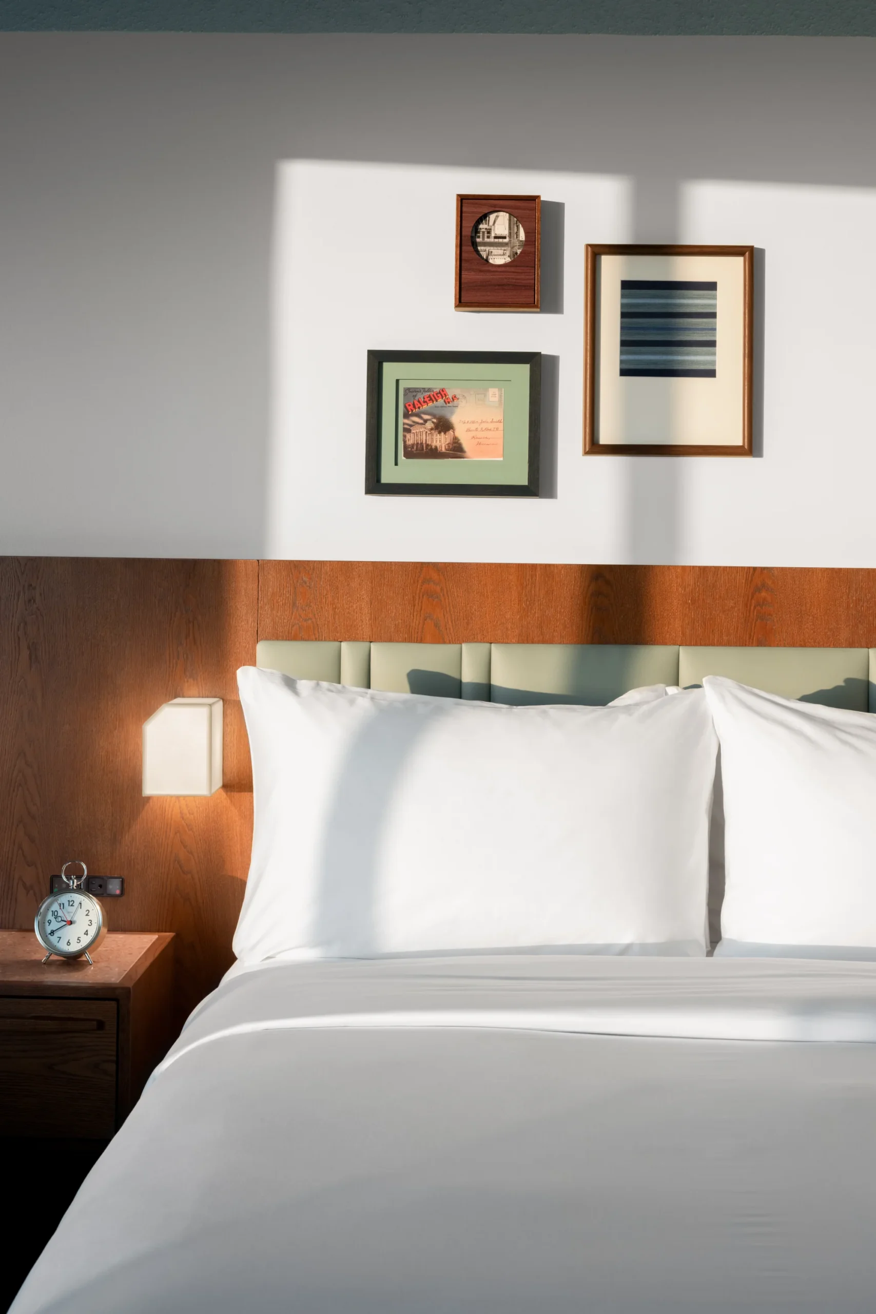 A bed with white sheets and pillows, a nightstand with an alarm clock, and two framed pictures on the wall.