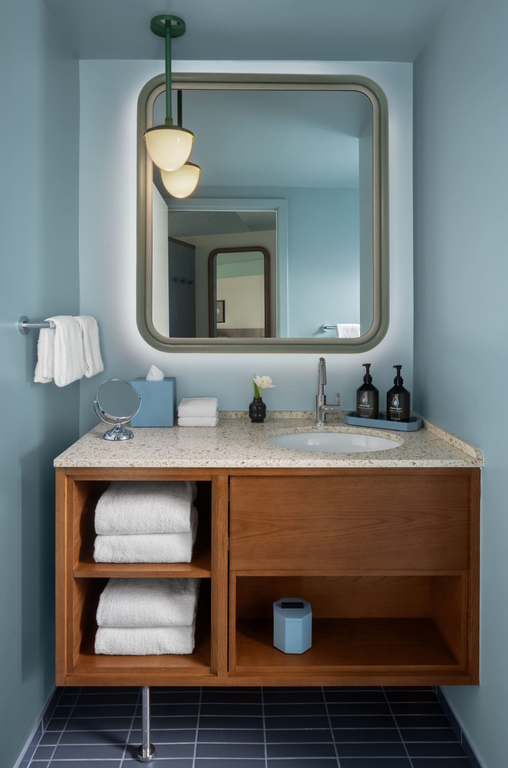 A bathroom vanity with a mirror, sink, and storage. The vanity features a marble countertop, a white porcelain sink, and a wooden cabinet with shelves and drawers. A white towel hangs on a towel rack to the left of the sink.