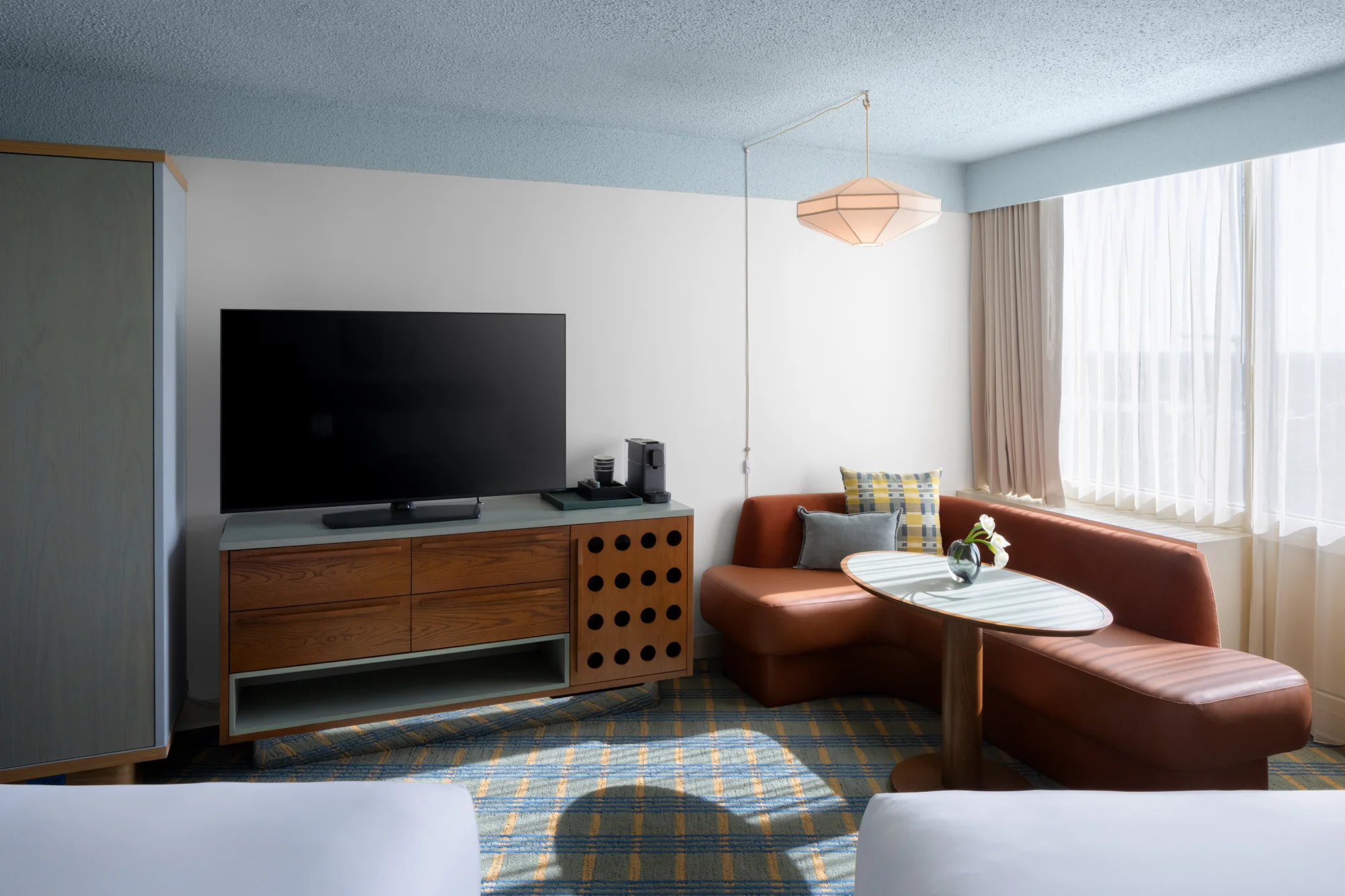 A hotel room with a TV, sofa, and table. The room is well-lit, with a large window on the right side. The sofa is red, and there is a small table in front of it. The TV is mounted on the wall, and there is a cabinet underneath it.