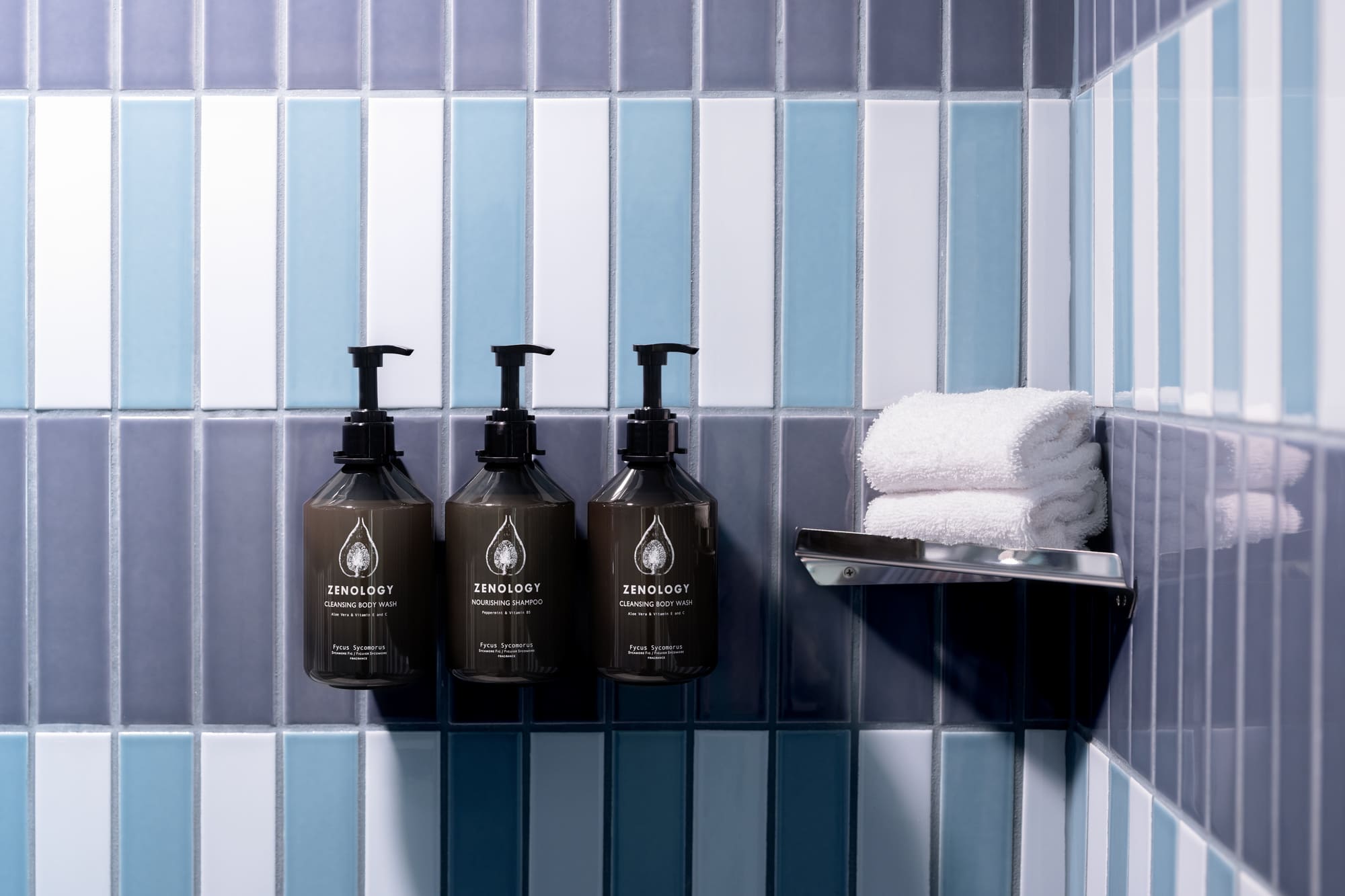 Two dark brown bottles of shampoo and conditioner with black pumps on a blue and white tiled shower wall, with a shelf holding a rolled-up white towel.