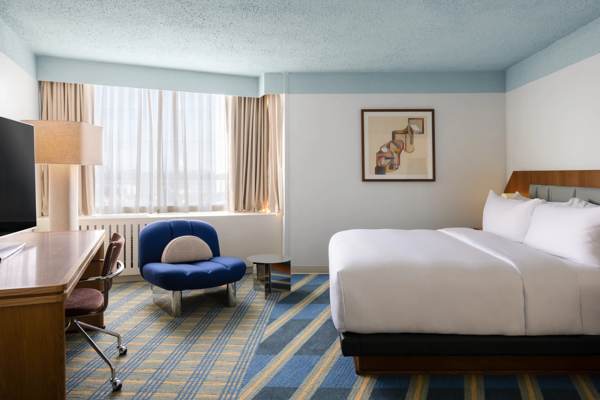 A hotel room with a large bed, blue chair, and desk, featuring a window with curtains and a picture on the wall.