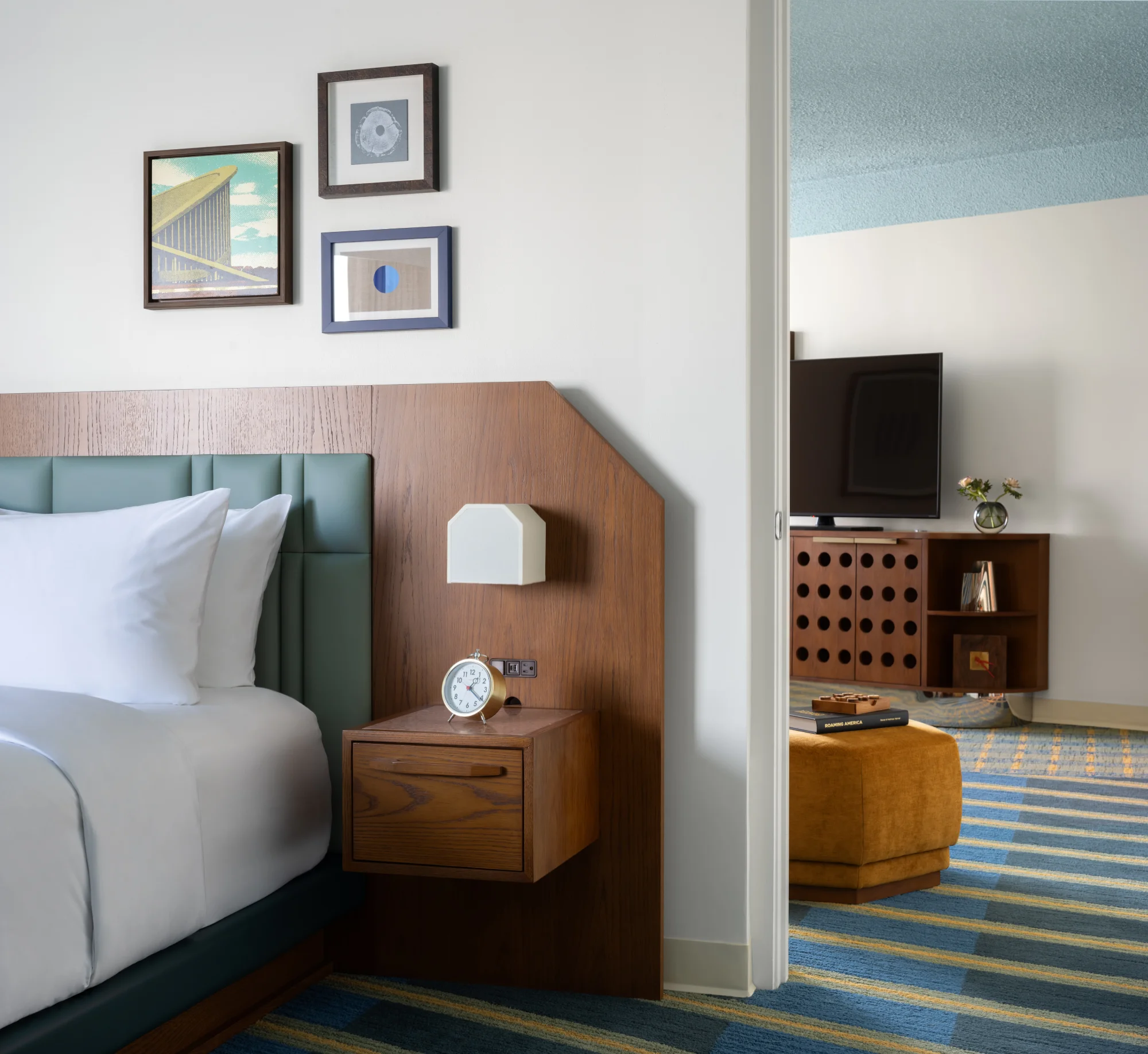 A bedroom with a bed, nightstand, TV stand, and striped rug. The bed has a white comforter and pillows, a wooden headboard, and a nightstand with a clock. The TV stand is in the background, and the rug has blue and yellow stripes.