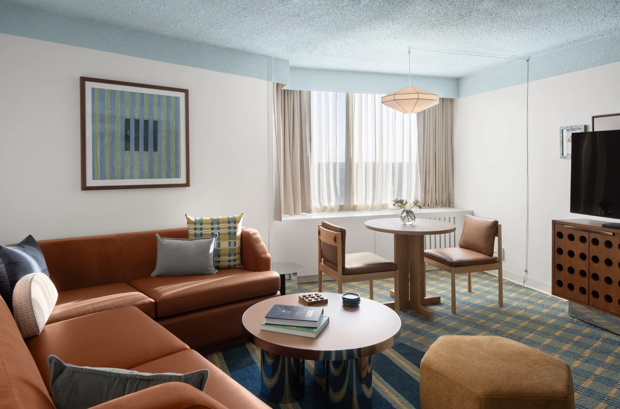 A living room with a brown couch, a coffee table, and a dining table. The room has a blue and yellow plaid carpet, a white wall with a picture, and a window with beige curtains.