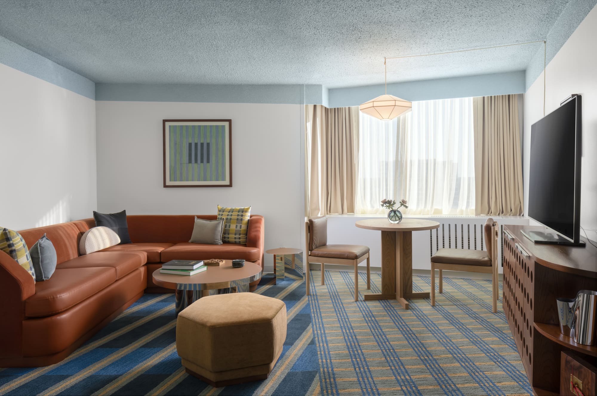 A living room with a couch, coffee table, and TV, featuring a blue and yellow striped rug and a window with beige curtains.