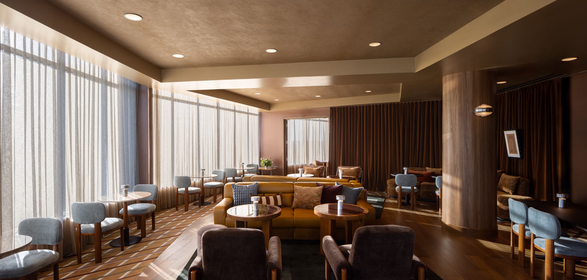 A dimly lit restaurant lounge with a large window on the left side, featuring a couch and chairs in the center, and a table and chairs on the right.