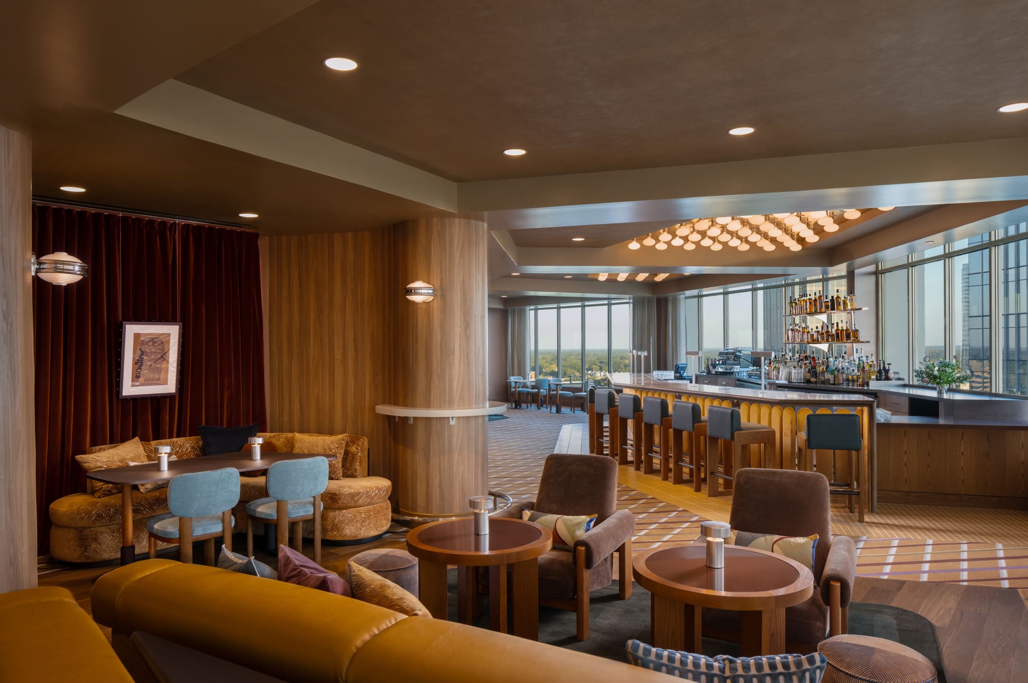 A spacious restaurant or bar with a large window, featuring a high ceiling, a bar with stools, and a seating area with tables and chairs.