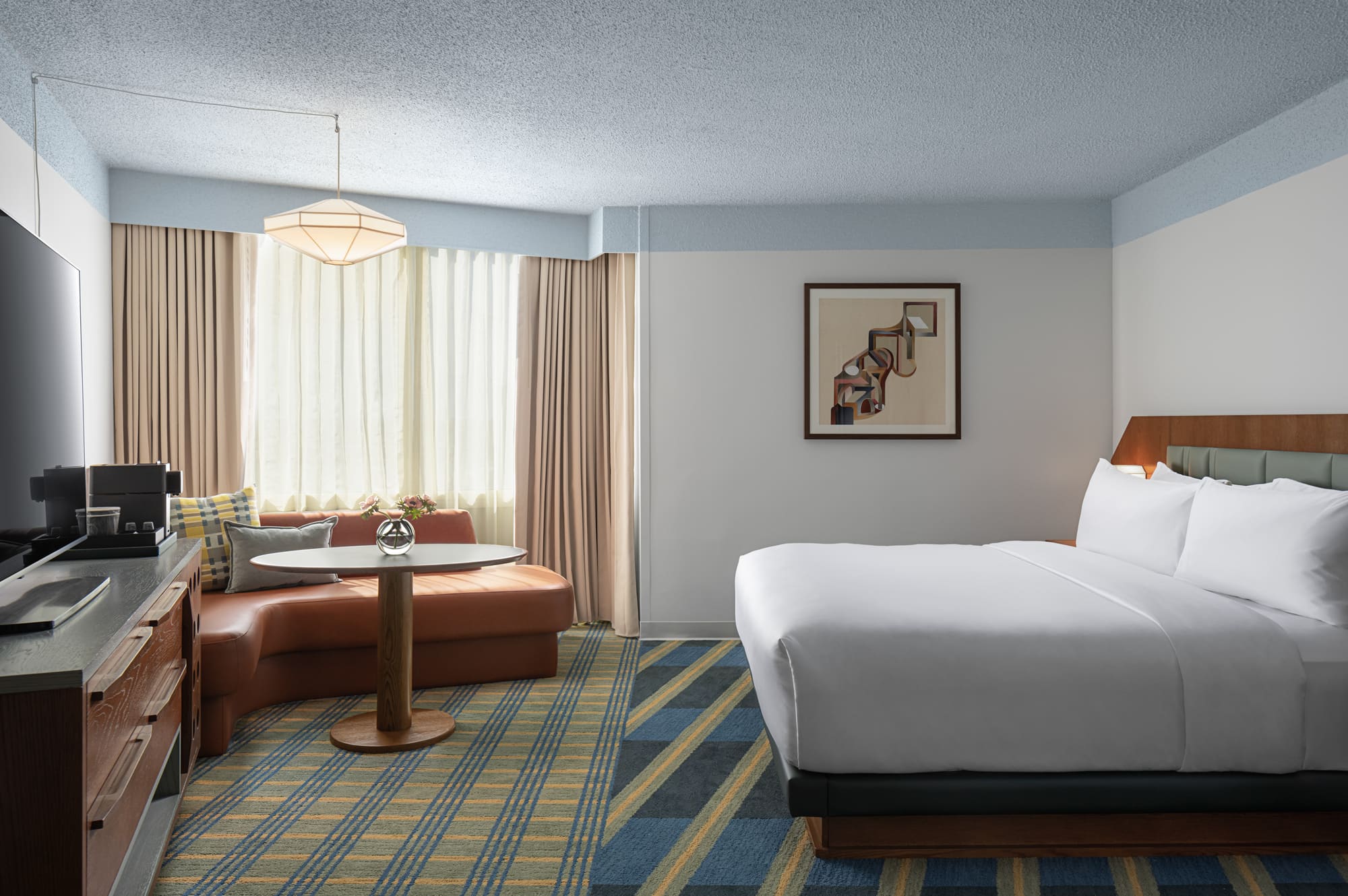 A hotel room with a king-sized bed, desk, and window. The room features a large bed with a white comforter and pillows, a small desk with a chair, and a window with beige curtains.