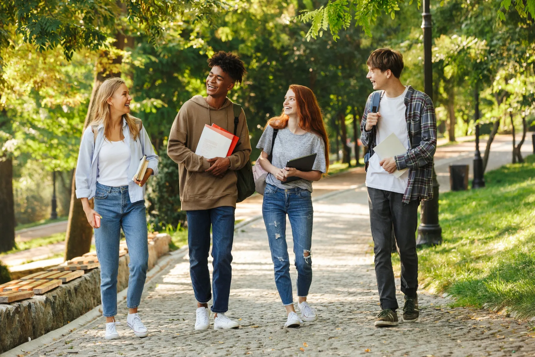Three young adults walking on a paved path in a park. The path is made of stone and has a low stone wall on the left side. The students are wearing casual clothing and carrying books and bags.