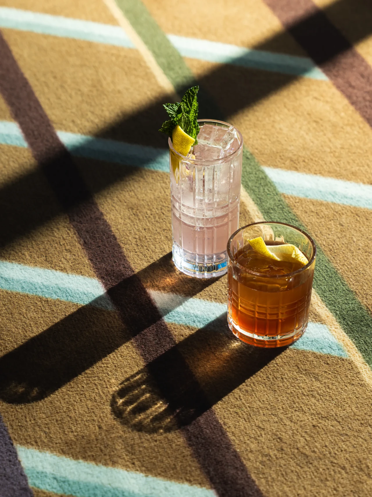 Two craft cocktails garnished with citrus and mint on sunlit plaid carpet