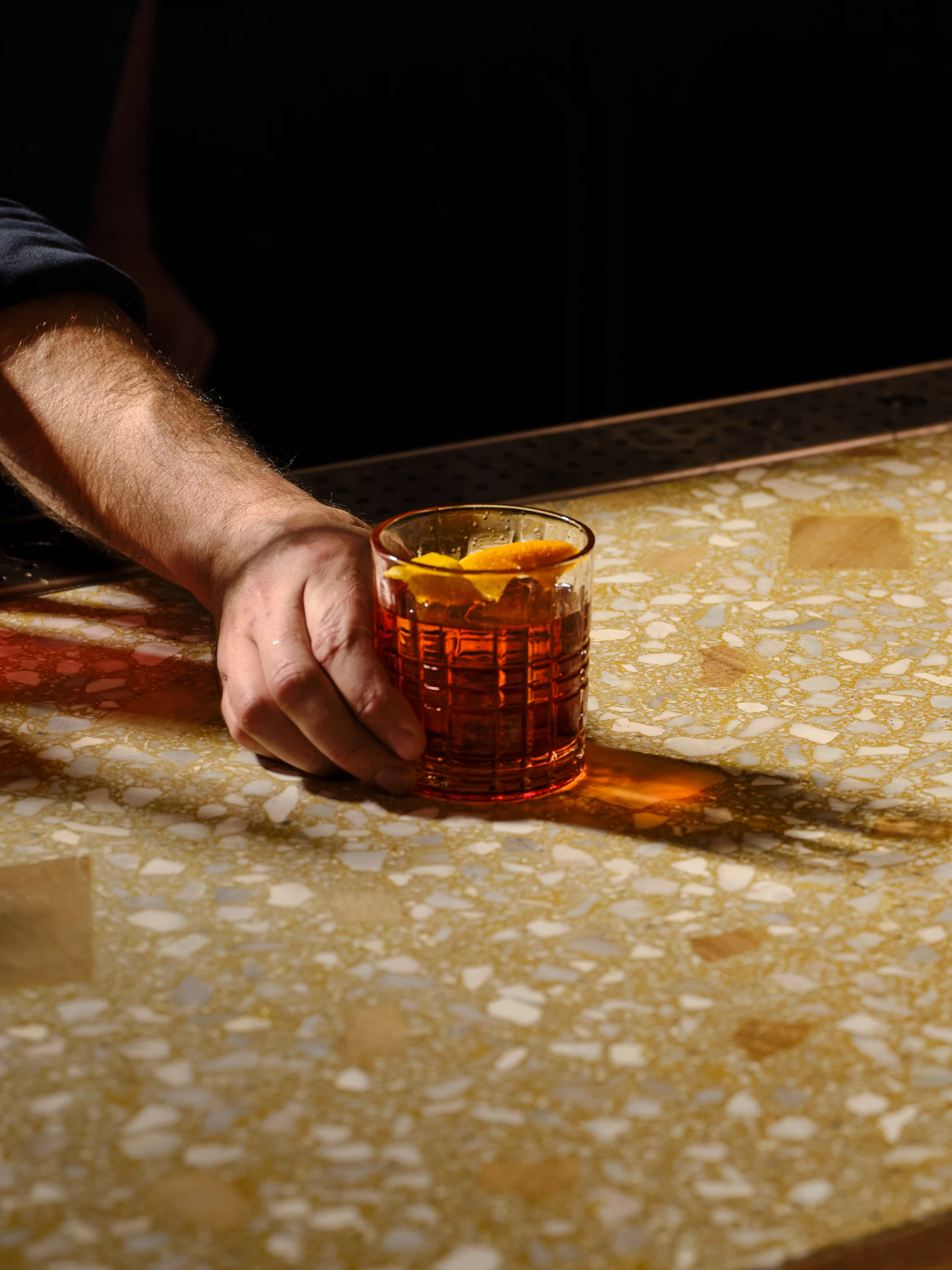 A bartender's hand sliding a negroni with an orange twist across the terrazzo bar at High Ground Lounge
